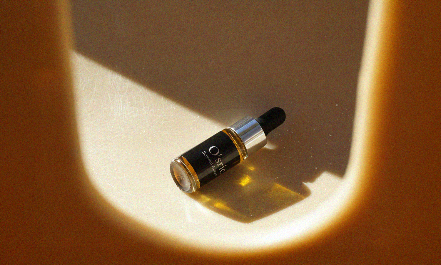 A bottle of O'sric scented essence oil on a beige surface with soft shadows